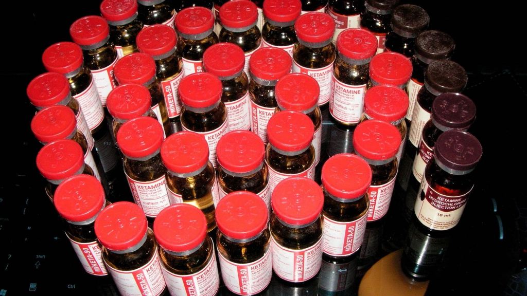 Rows of small glass vials of ketamine hydrochloride injection with red flip-off caps, standing together on a dark glossy surface