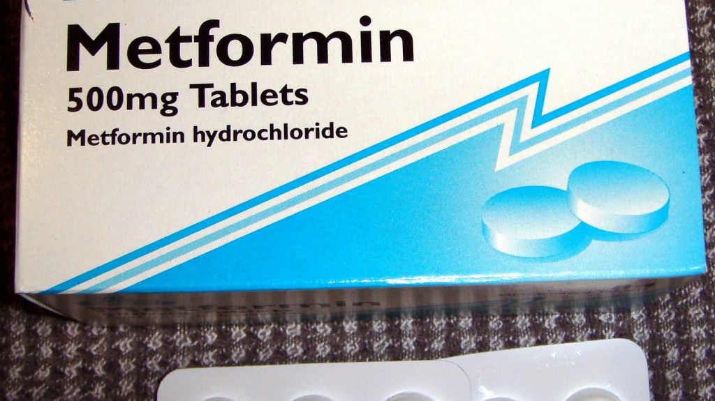 A white and blue box of Metformin 500 mg tablets beside a blister strip of round white pills on a grey carpet