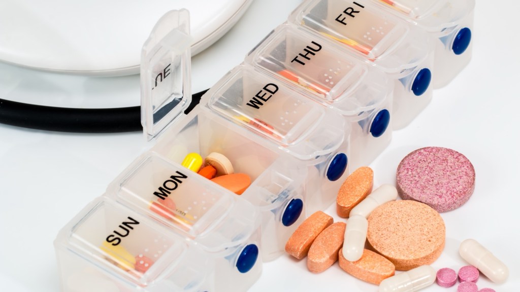A clear plastic weekly pill organizer labelled Sun to Sat on a white table, the Monday lid open, with orange vitamin tablets, white capsules and pink tablets scattered beside it