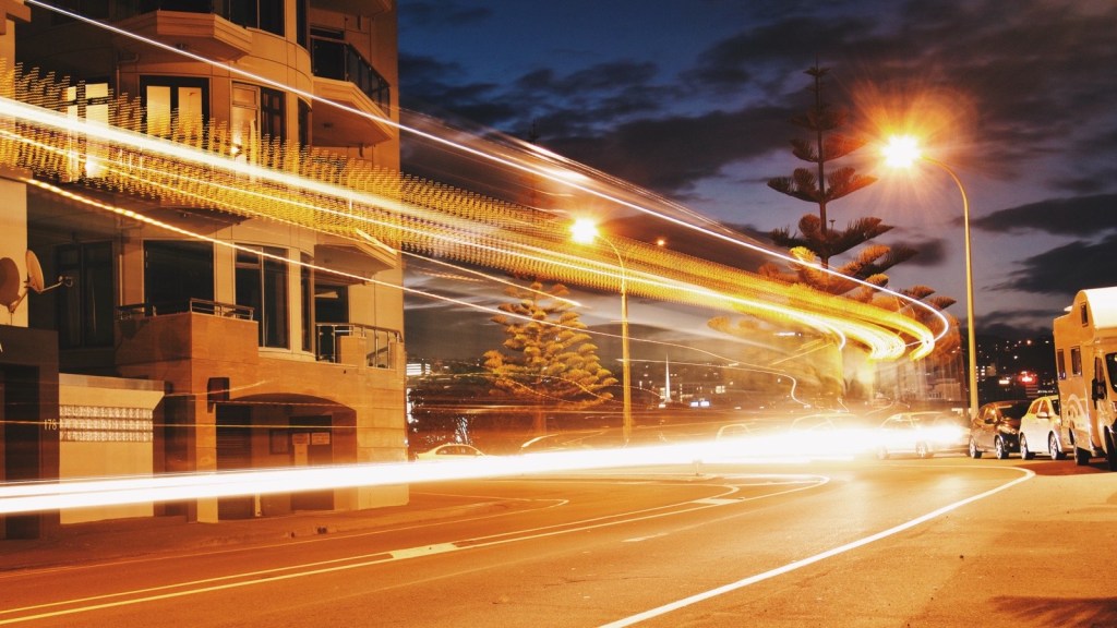 Streaks of car headlights curving along a road at dusk, past an apartment building with balconies, under orange streetlights and a dark cloudy sky