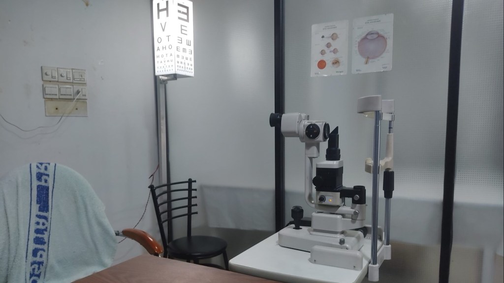 An eye exam room with a slit lamp microscope on a white table, an illuminated letter chart on a stand and anatomy posters of the eye on a frosted glass wall