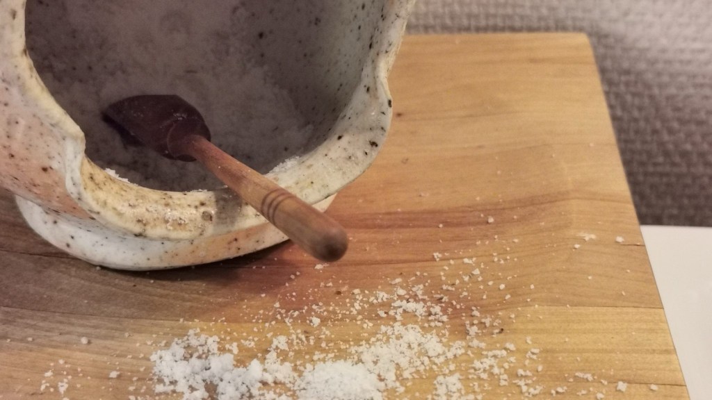A speckled ceramic salt cellar with a small wooden spoon on a wooden board, with coarse white salt spilled across the wood