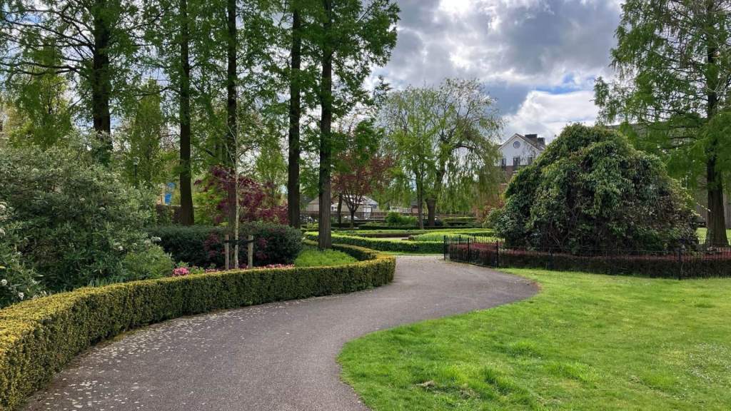 A paved path curving through a quiet green park, between a trimmed hedge and a lawn, under tall trees and a cloudy sky