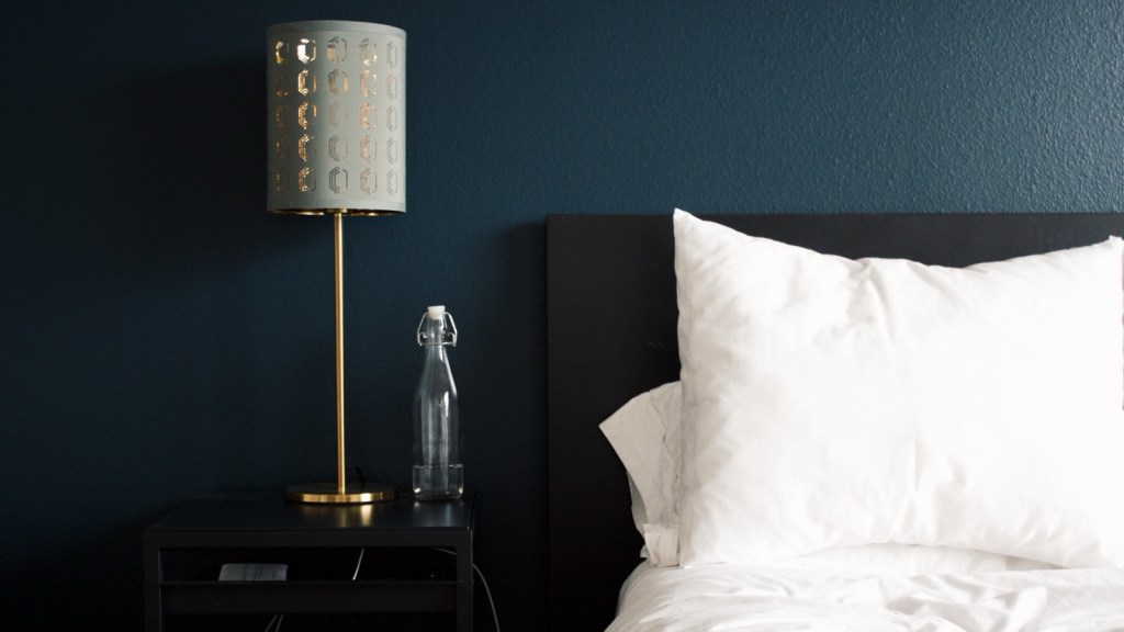 A brass bedside lamp and a glass water bottle on a black nightstand beside a bed with white pillows, against a dark navy bedroom wall