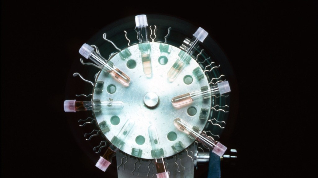 Small capped glass tubes of pink cell-culture liquid clipped around a spinning metal wheel, lit against a black background